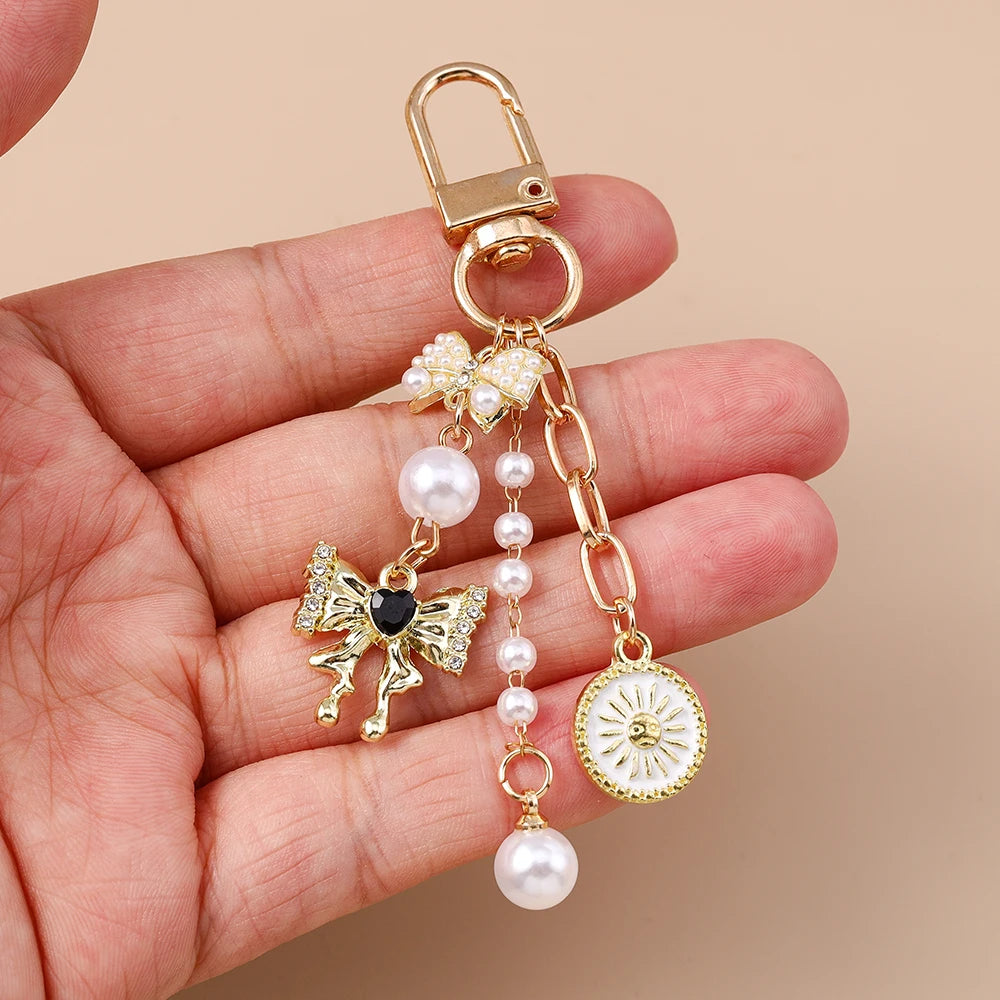 A gold-toned pearl bow heart keychain with a dangling charm is held in a person's hand.