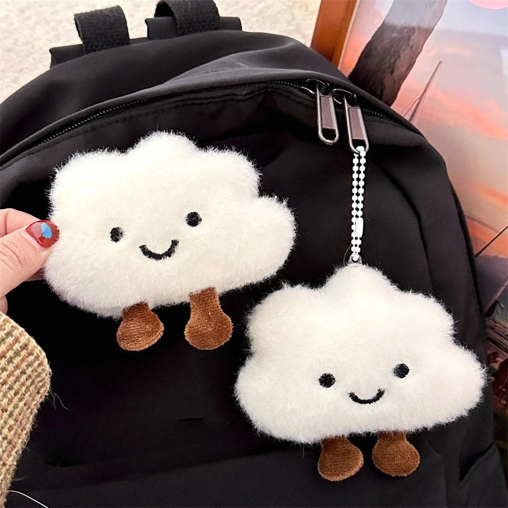 A white cloud shaped plush keychain with brown feet is attached to a black backpack.