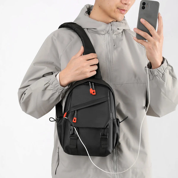 A man wearing a gray jacket is holding a black outdoor sports chest bag and a white charging cable.