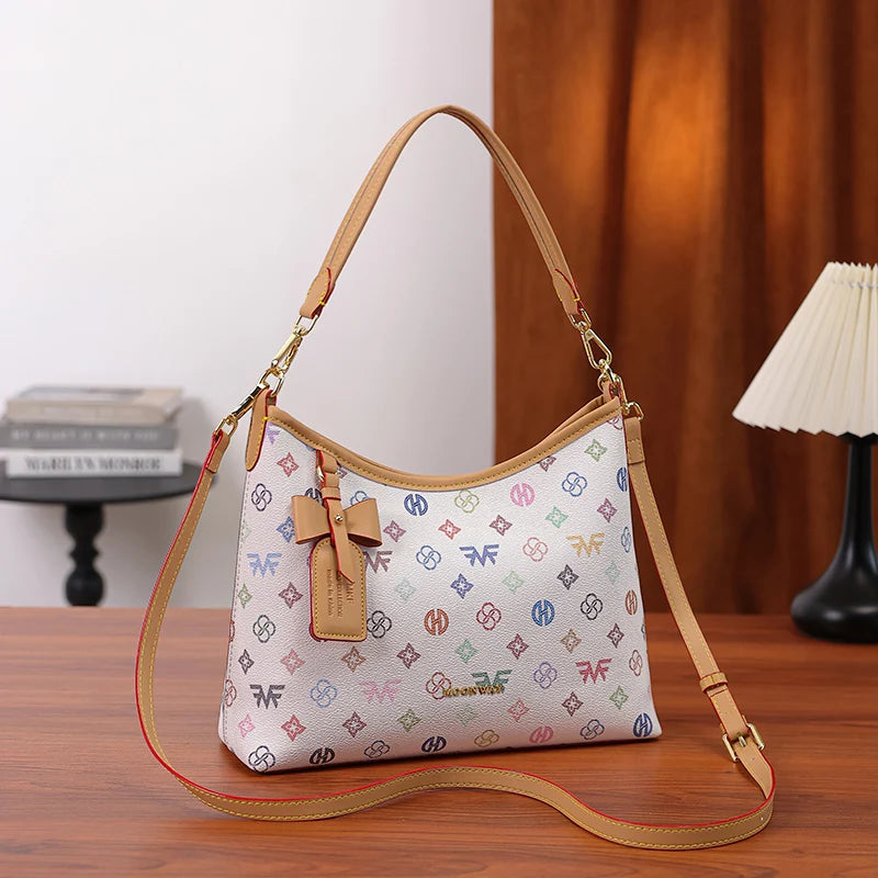 A beige crescent tote bag with a letter print and bow accent resting on a wooden surface.