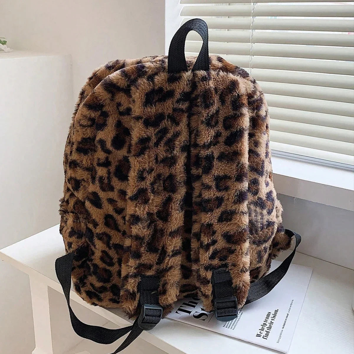 A brown leopard print plush backpack hanging on a white shelf next to a window.