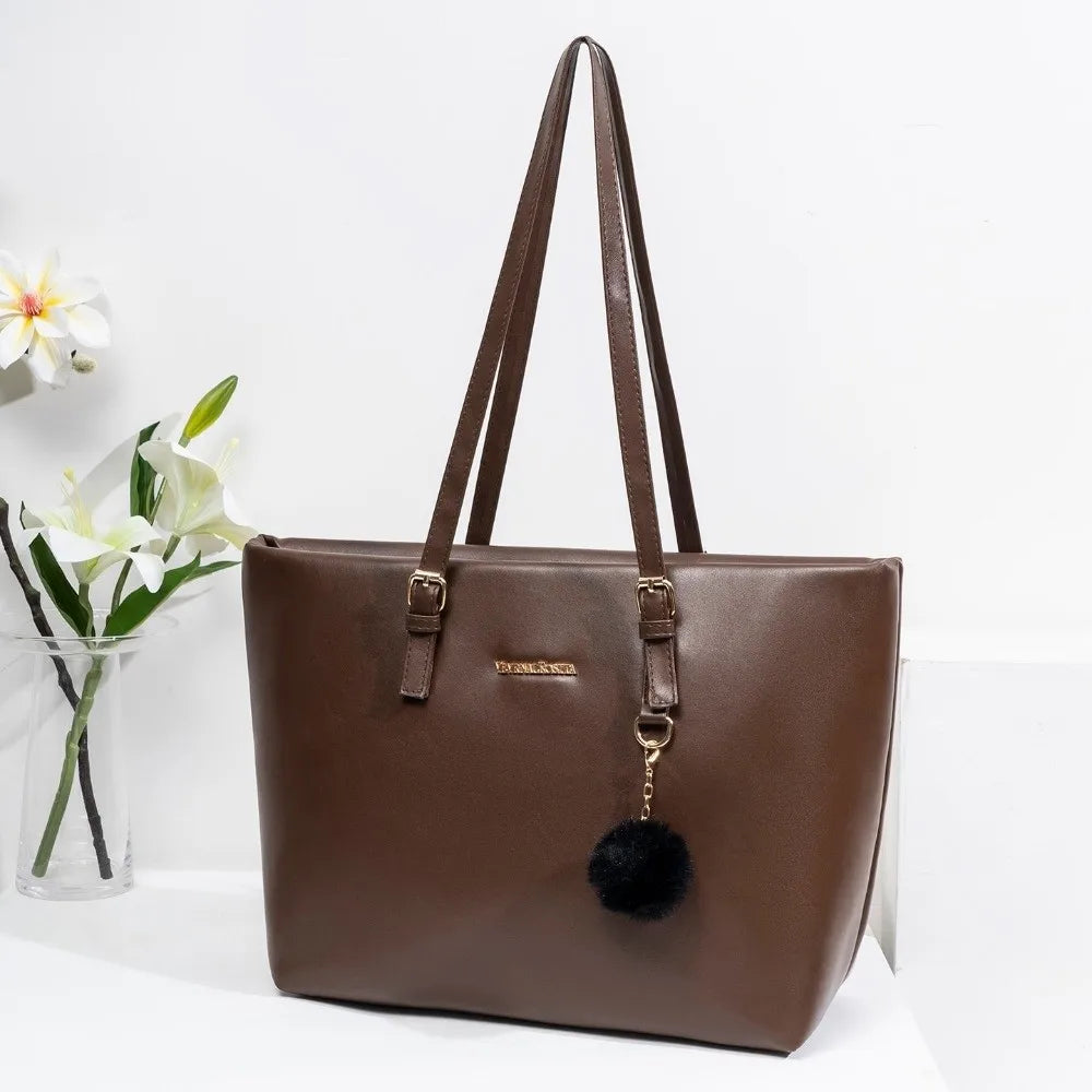 Bolso de hombro de cuero PU marrón con dije dorado y pompon negro, exhibido sobre una superficie blanca.