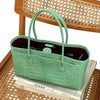 A light green crocodile print leather handbag resting on a stack of books, set against a woven wooden background.
