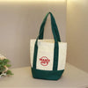 A white canvas bag with dark green handles and trim, displayed on a light surface.