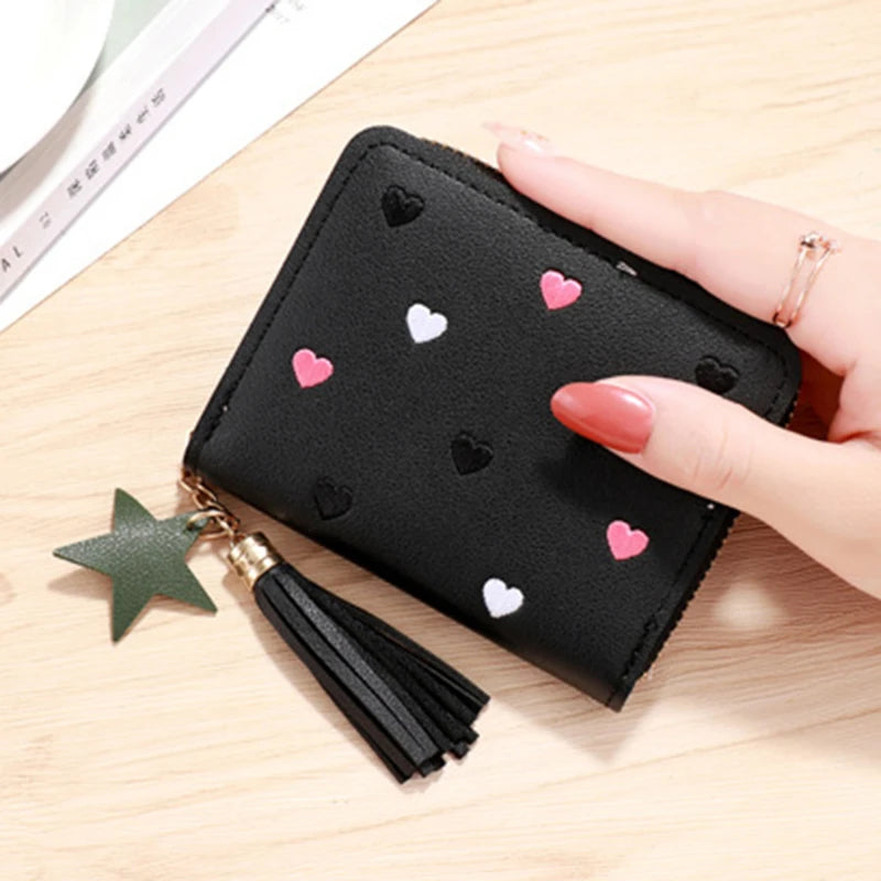 A black PU leather wallet with a zipper closure decorated with small pink and white heart patterns, shown next to a gold tass