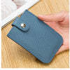 A hand holds a blue-gray genuine leather slim card holder with a silver stud closure on a wooden surface.