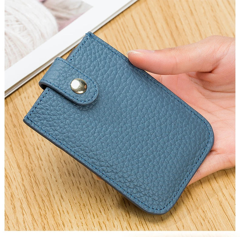 A hand holds a blue-gray genuine leather slim card holder with a silver stud closure on a wooden surface.