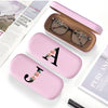 A pink PU leather hard shell eyeglass case with a gold letter charm resting on a white background.