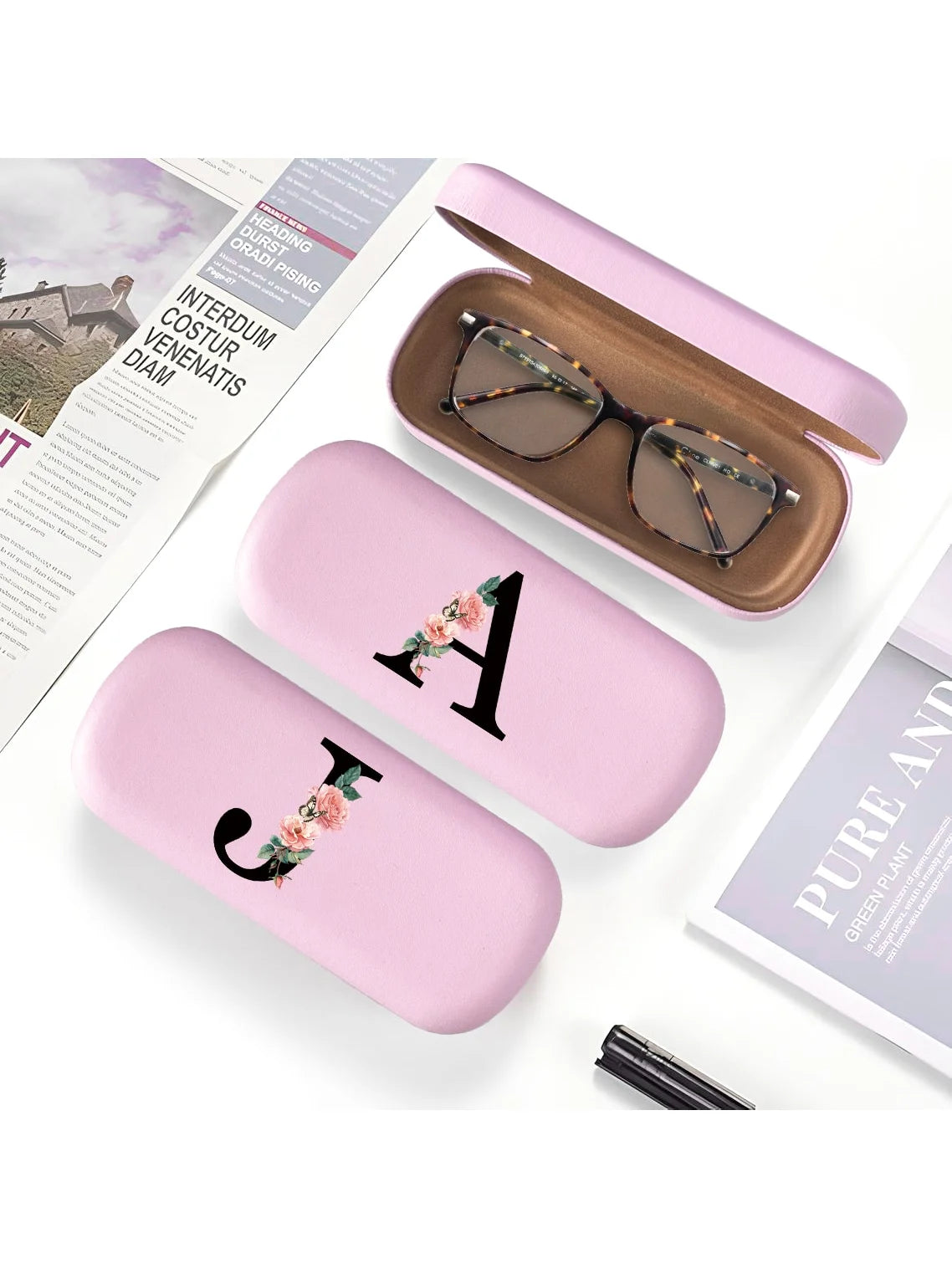A pink PU leather hard shell eyeglass case with a gold letter charm resting on a white background.