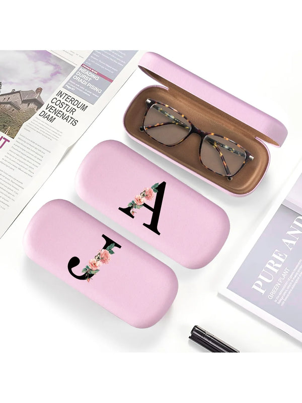 A pink PU leather hard shell eyeglass case with a gold letter charm resting on a white background.