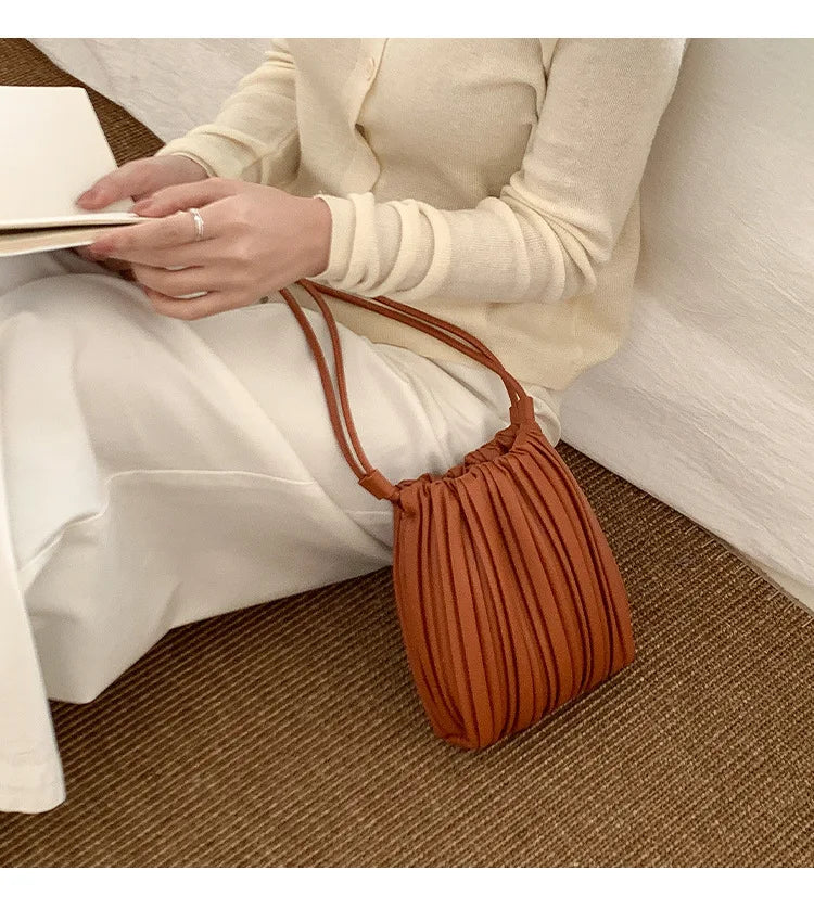 A woman holds a pleated brown PU leather crossbody bag while sitting on a floor.