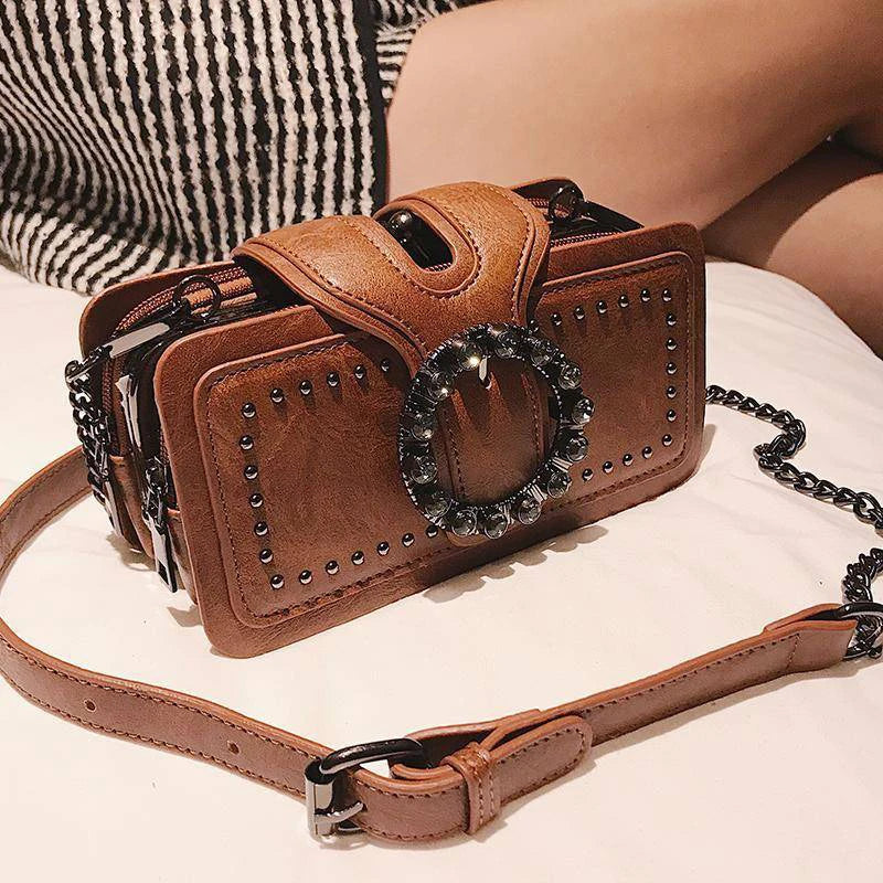 A brown PU leather crossbody bag with a studded design and a rhinestone buckle resting on a white surface.