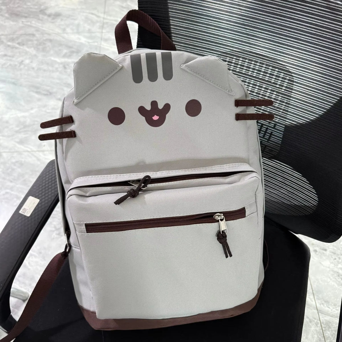 A light gray Pusheen cat canvas backpack with brown accents resting on a dark chair.