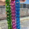 A hand is holding a thick, colorful acrylic Cuban link necklace featuring green, pink, and blue links against a bright sky ba