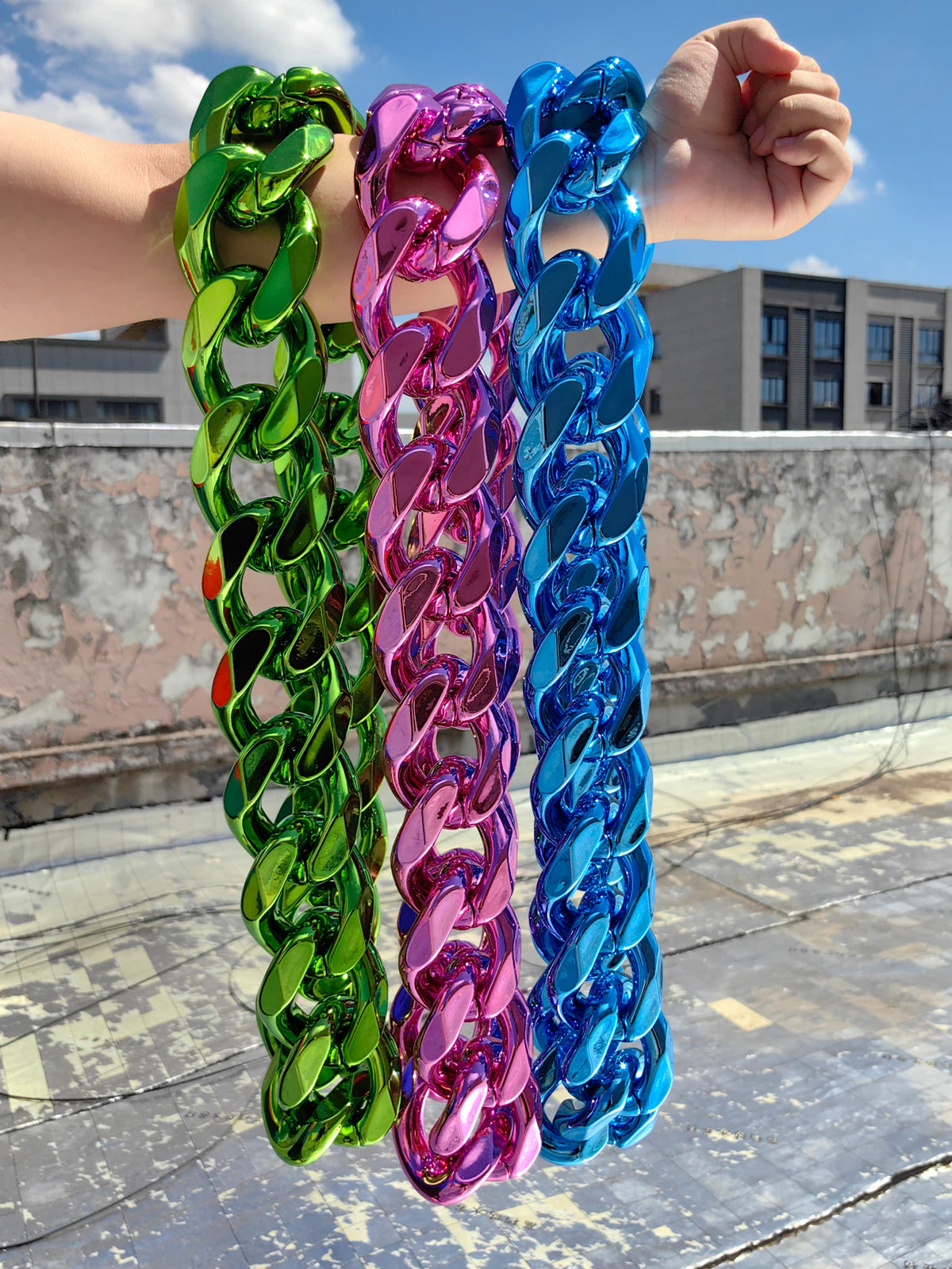 A hand is holding a thick, colorful acrylic Cuban link necklace featuring green, pink, and blue links against a bright sky ba