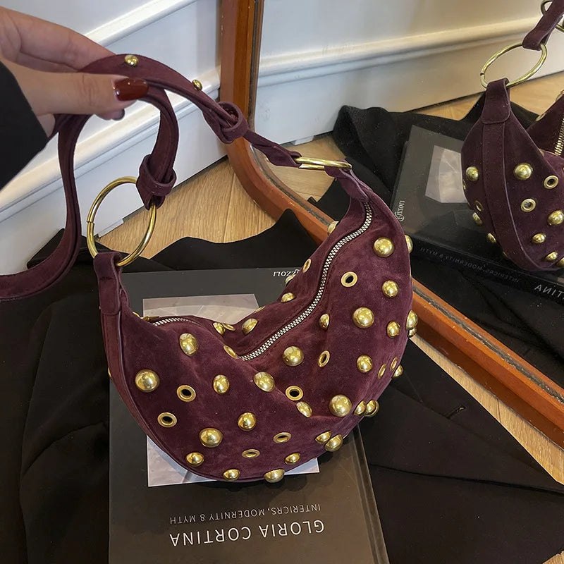 A dark purple faux suede hobo crossbody bag with gold rivet accents displayed on a wooden surface.