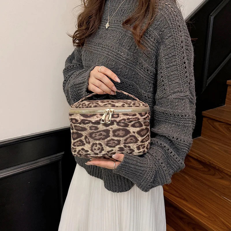 A woman wearing a gray sweater holds a brown faux suede cosmetic bag in front of a dark staircase.