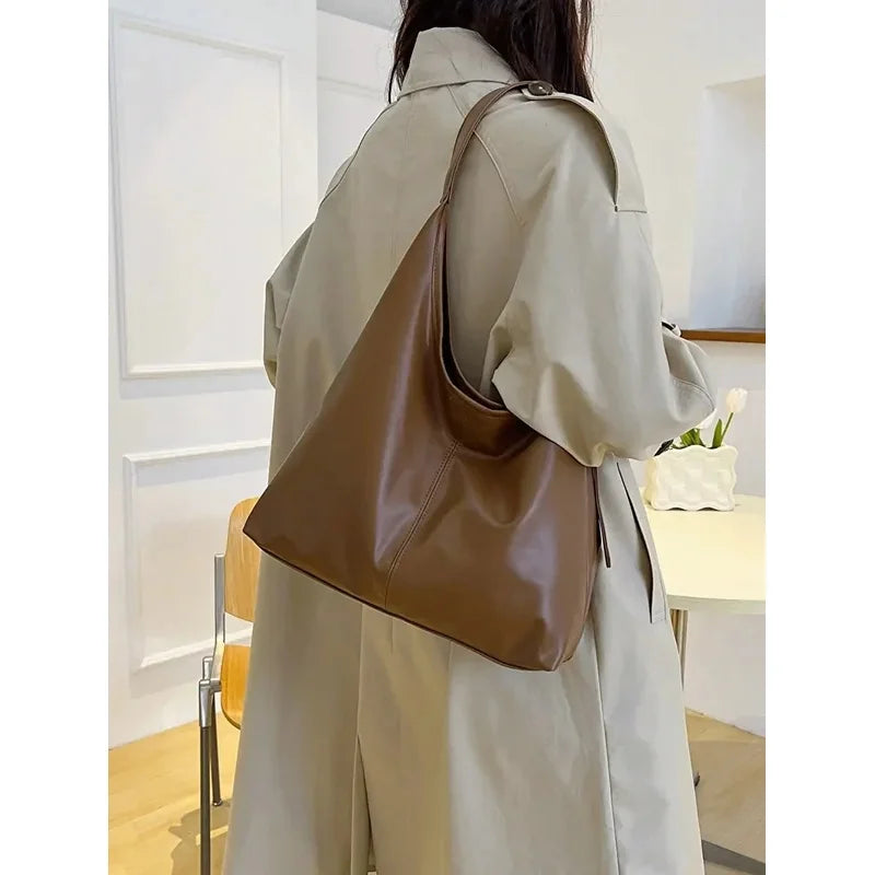 A woman is shown from behind wearing a beige trench coat and carrying a brown PU leather slouchy hobo shoulder bag.