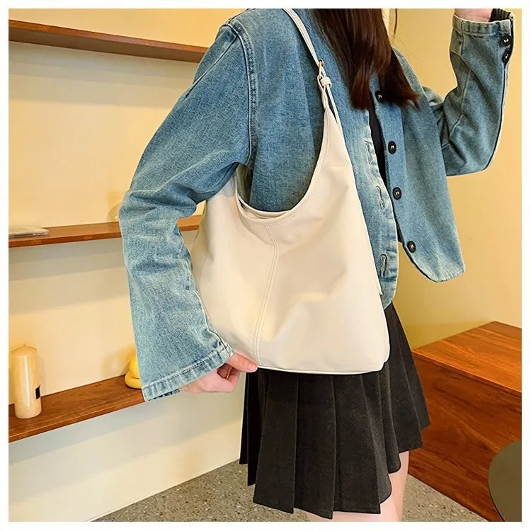 Eine Frau hält eine cremefarbene Leinencoffertasche über einem schwarzen plissierten Rock und trägt eine Jeansjacke.