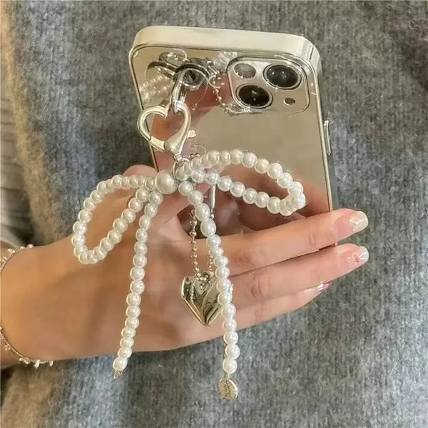 A hand holds a pearl bowknot keychain with a heart charm against a gray sweater.