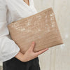 A beige textured clutch bag with a subtle alligator print is held by a person wearing a white shirt.