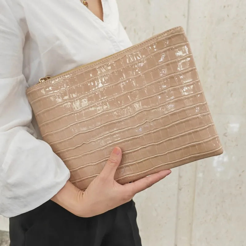 A beige textured clutch bag with a subtle alligator print is held by a person wearing a white shirt.