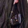 A black genuine leather square shoulder crossbody bag is held by a hand against a dark fabric background.