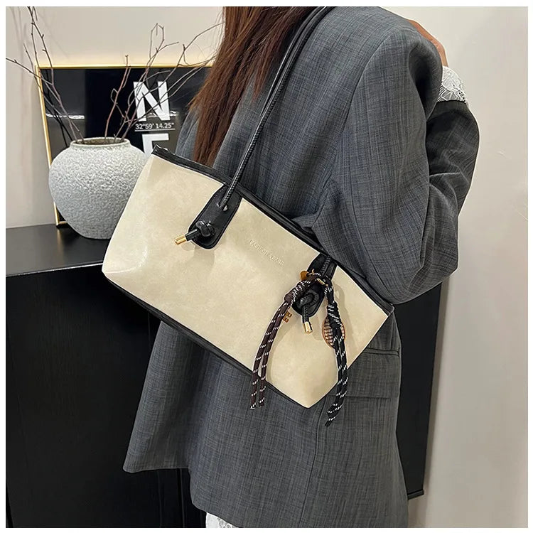 A woman holds a beige classic PU leather tote bag with black leather strap and gold hardware against a gray blazer.