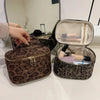 A brown faux suede cosmetic bag with a gold zipper is shown open, revealing makeup products and a mirror in the background.