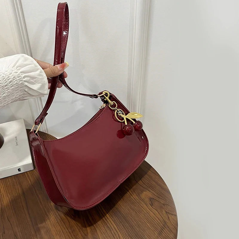 A maroon PU leather shoulder bag with a strap and gold cherry pendants is being held by a hand.