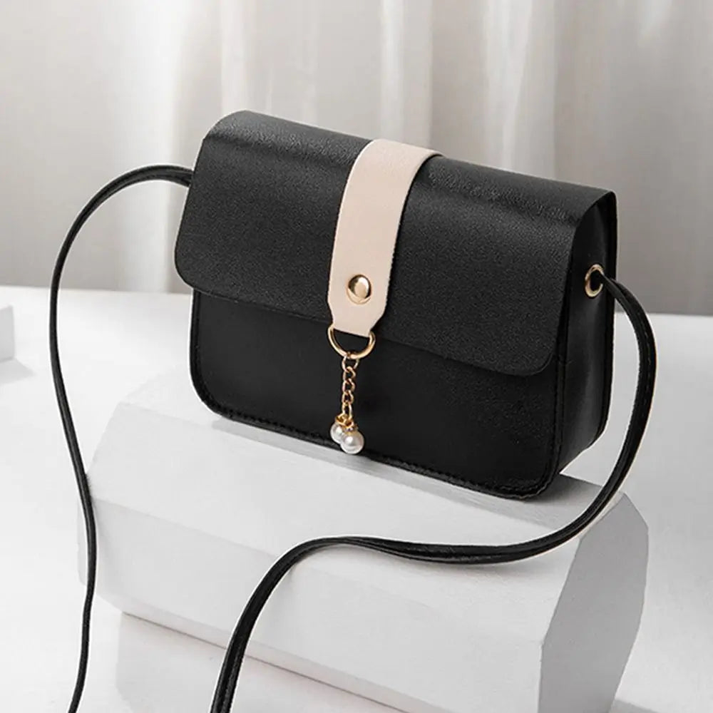 A black fashion square crossbody bag with a light beige strap and gold chain detail resting on a white surface.
