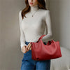A woman wearing a beige turtleneck sweater and blue jeans holds a red leather tote bag.