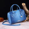 A light blue crocodile pattern luxury leather crossbody tote bag resting on a pink surface next to a piece of driftwood.