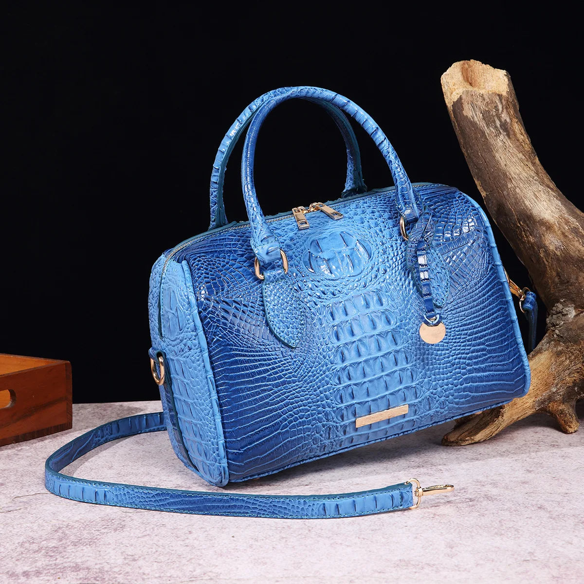 A light blue crocodile pattern luxury leather crossbody tote bag resting on a pink surface next to a piece of driftwood.