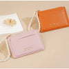 Hellorange Leder-Kartenwallet mit goldener Reißverschluss und Perlenkette neben einer rosa Ledergeldbörse.