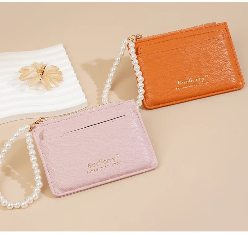 Hellorange Leder-Kartenwallet mit goldener Reißverschluss und Perlenkette neben einer rosa Ledergeldbörse.