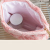 A pink plush storage bag lined with pink fabric holds a round cosmetic container on a metal tray.