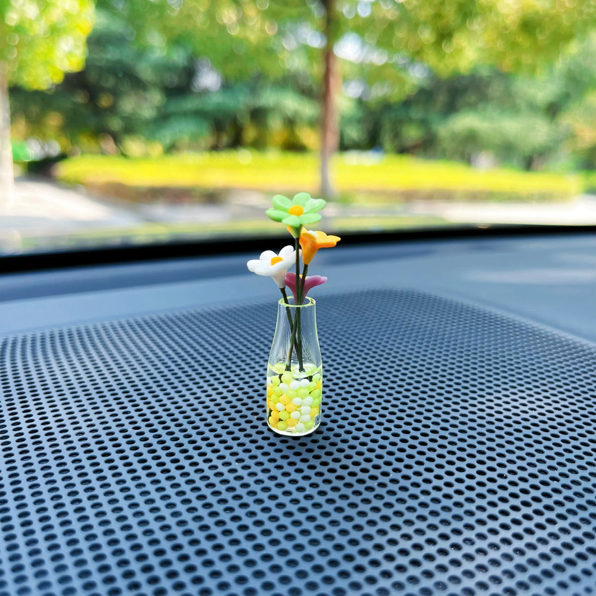 Ein Glasvase mit farbenfrohen Blütenblättern und kleinen gelben Kugeln auf einer dunklen Auto-Innenausstattung.