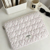 A quilted white laptop sleeve with a pink floral pattern rests on a white table next to a mug and a box of books.