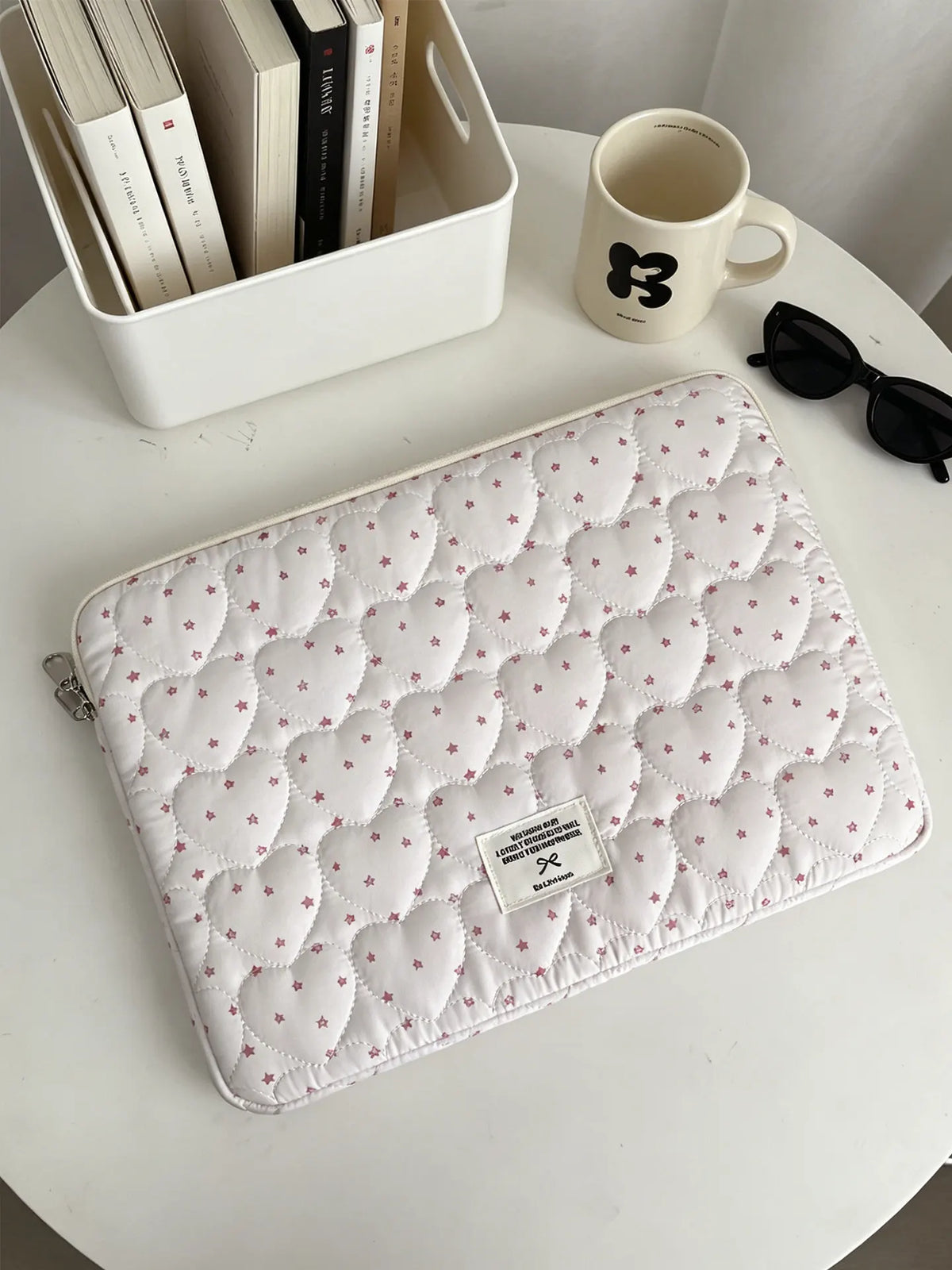A quilted white laptop sleeve with a pink floral pattern rests on a white table next to a mug and a box of books.