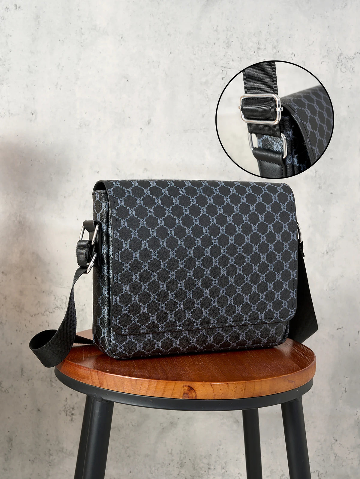 A black patterned flap crossbody bag is displayed on a wooden stool with a close-up of the strap detail.