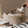 A hand holds a beige foldable cosmetic storage bag with makeup brushes inside, resting on a white cloth.
