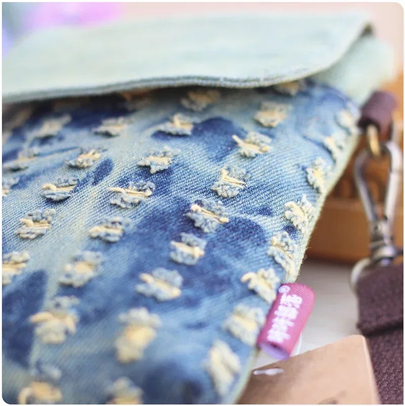 A close-up of a denim mini messenger bag with light blue fabric and yellow stitching, showing the texture and detail.