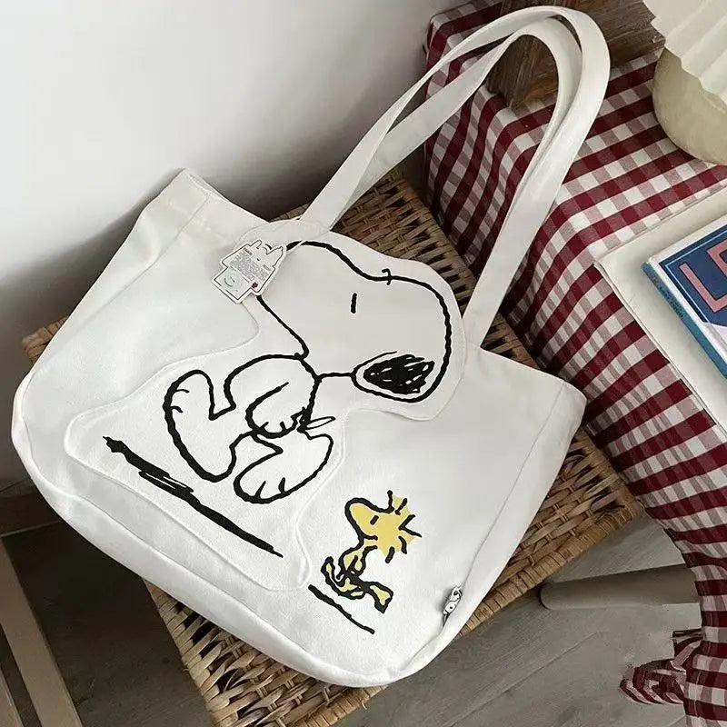 A white canvas shoulder bag featuring a Snoopy cartoon design, resting on a woven mat.