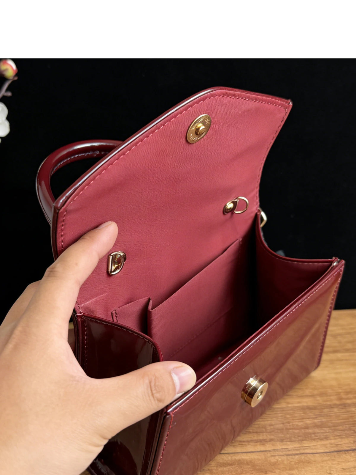 A hand opens a maroon glossy texture evening handbag, showing the interior lining and hardware.