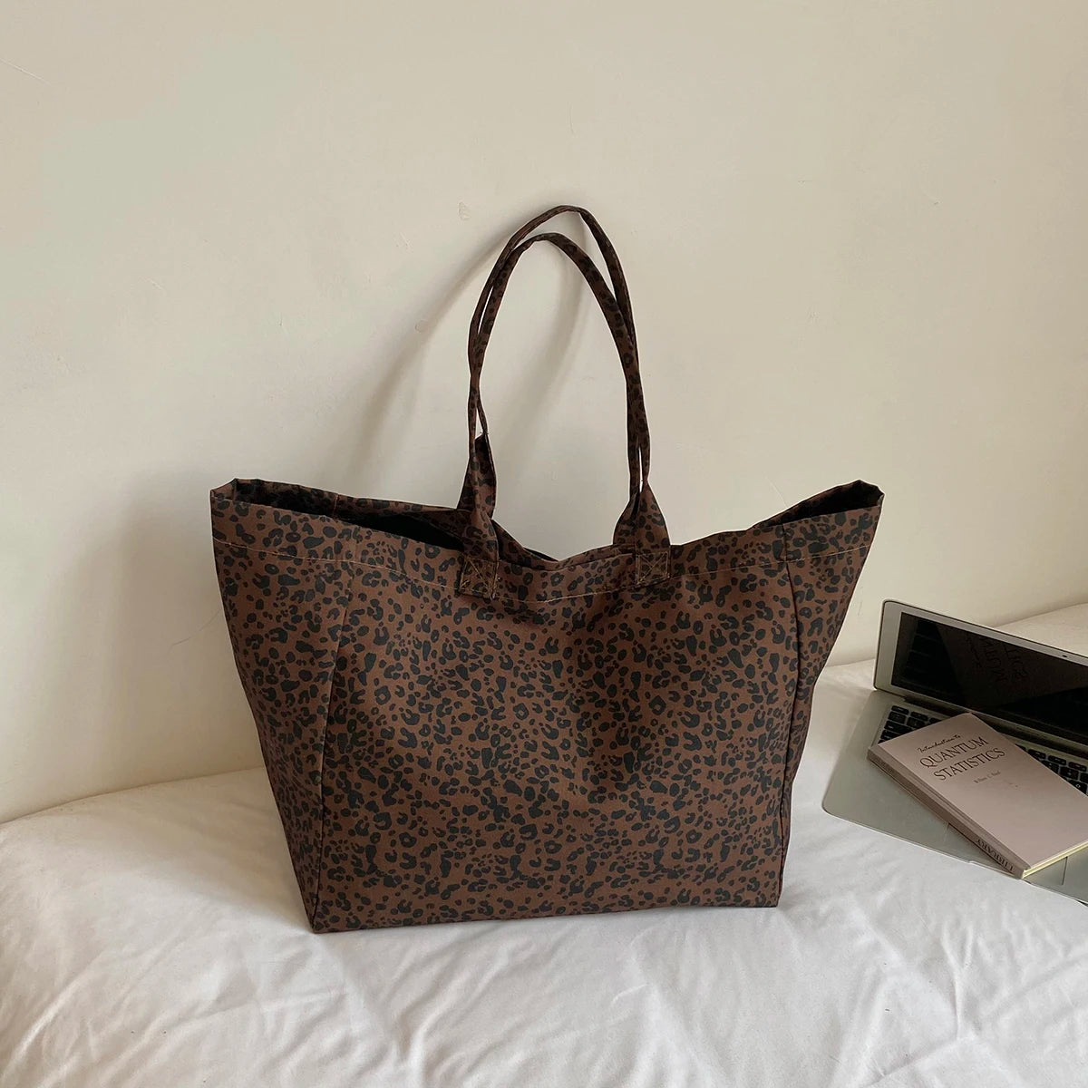 A brown leopard print canvas tote bag with handles resting on a white surface next to a notebook.