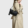 A woman wearing a beige trench coat holds a black split leather shoulder tote bag.