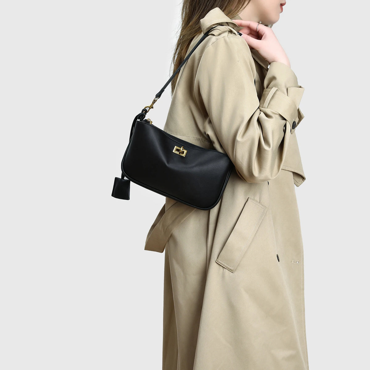 A woman wearing a beige trench coat holds a black split leather shoulder tote bag.