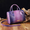 A purple and blue crocodile pattern luxury handbag resting on a wooden surface next to a piece of driftwood.