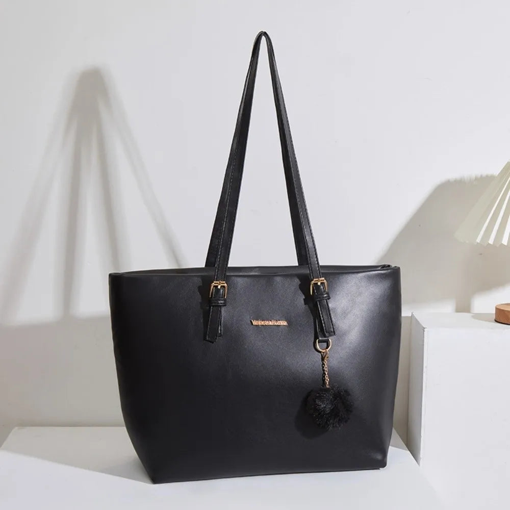 Bolso tote de cuero PU negro con herrajes dorados y un adorno de pompon negro sobre una superficie blanca.